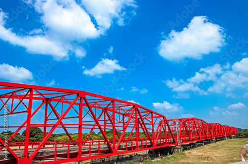 A striking red steel truss bridge stretching gracefully across the landscape, its vivid color contrasting with the serene greenery and bright sky.
