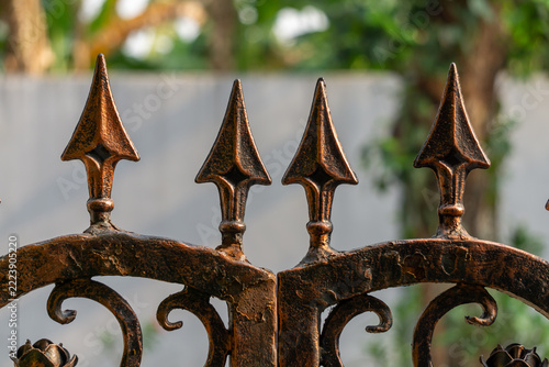 wrought iron fence. Rusted iron spikes stand tall and sharp. Curved metalwork frames the pointed tips. Weathered surface hints at age and exposure. Faded greenery blurs softly in the background