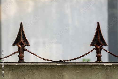 Rusted metal spikes guard a chain-link fence. Weathered iron points pierce the gray background. Chain links stretch between ornate posts. Faded steel tones suggest aged durability