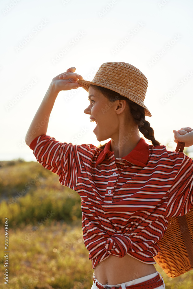 Fototapeta premium Young woman enjoying summer lifestyle outdoors. She wears a striped shirt and straw hat, holding a basket. Sunset light, concept for vacation, freedom, leisure and joy.