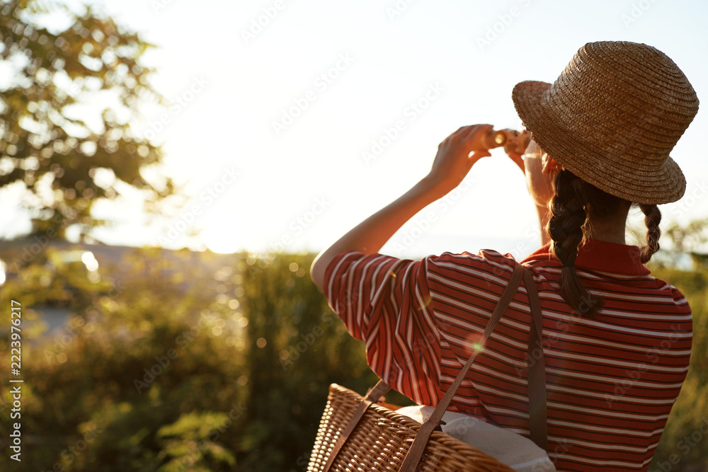 Fototapeta premium Young woman in hat taking pictures with camera outdoor. Lifestyle portrait with summer sun flare. Concept of travel, photography, hobby, and relaxation in nature.