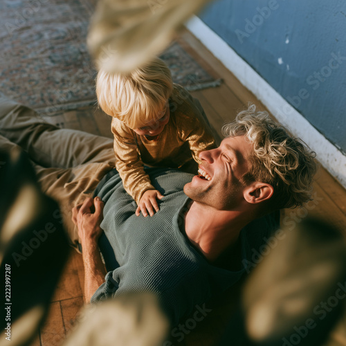 Sunlight bathes a father and child in tender warmth. Child rests atop father's chest, smiling wide. Father lies back, eyes closed, laughing softly. Their bond shines through quiet joy and comfort