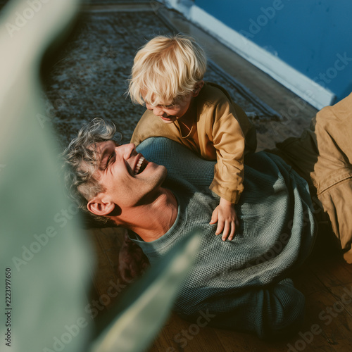 Man laughs joyfully as child sits on his chest. Soft sunlight bathes the wooden floor and blue wall. Child's golden hair and bright eyes show delight. Warm, candid moment captures pure familial love