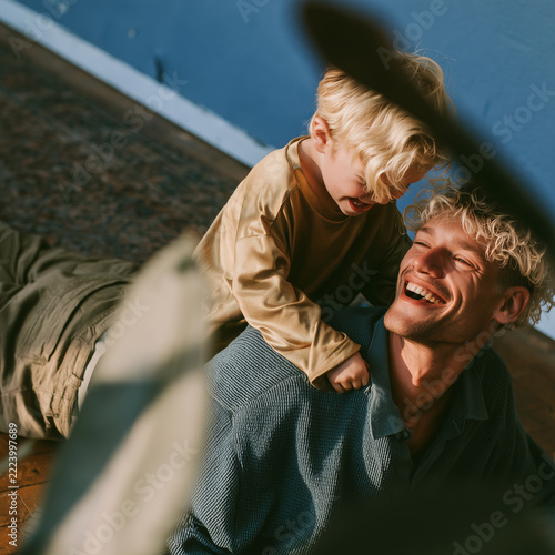 Sunlight glows warmly on a father and child. Child smiles, resting on father's shoulders. Father laughs, eyes closed, fully present. Their joy radiates through shared affection