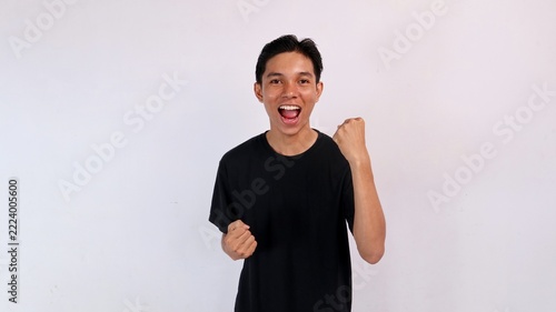 Cheerful young Asian man smiling with an open mouth while raising his left fist in celebration against a white studio background.