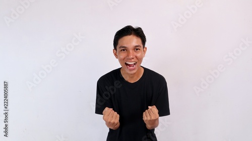Joyful young Asian man shouting happily with mouth open while holding both clenched fists to celebrate an achievement against a white background.