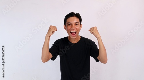 Ecstatic young Asian man shouting with joy while raising both fists high in victory against a clean white background.