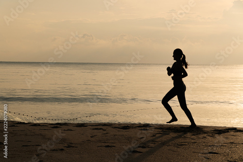 Obraz na plátně Silhouette of a young active woman running barefoot on a tranquil beach at golden sunrise, promoting health, fitness, and an energetic lifestyle with ocean background copy space