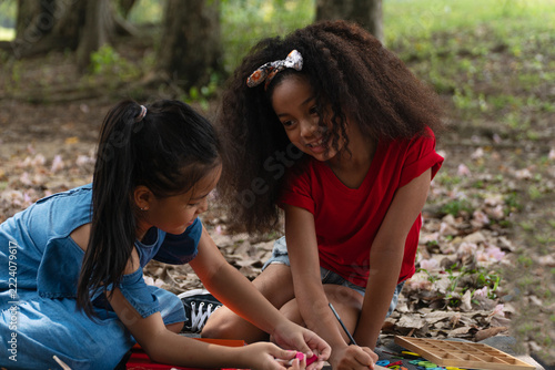 Two happy diverse little girls sitting together in an outdoor park, smiling, drawing, and playing with educational wooden alphabet toys, representing childhood friendship, teamwork, and learning.