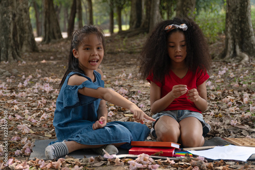 Two diverse little girls sitting on a mat in an outdoor park with books and drawing paper, one happy Asian girl pointing a finger while playing and learning together in childhood friendship.