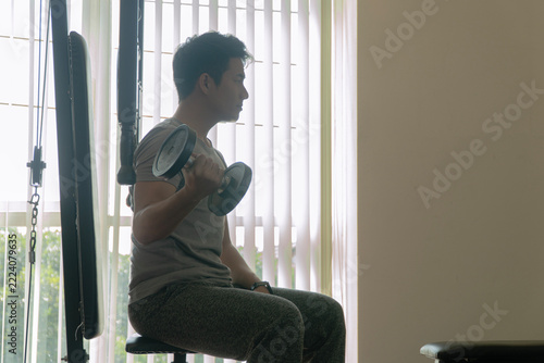 Profile view of a determined young Asian man sitting on a bench in a gym and lifting a dumbbell, focusing on bicep curls, representing strength training, fitness motivation, and an active lifestyle.