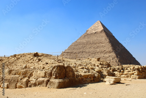 The Great Pyramid of Khafre in Giza standing tall under a clear blue sky, viewed over ancient stone ruins and desert landscape in Egypt.