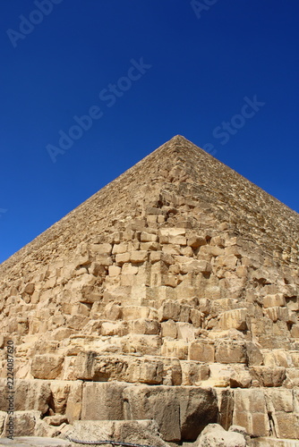​A low-angle, vertical close-up shot showing the textured limestone blocks and imposing structure of the Great Pyramid of Giza against a clear blue sky.