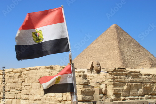 Egyptian national flags waving in the foreground with the Great Sphinx and the Pyramid of Khafre rising in the background under a bright blue sky.