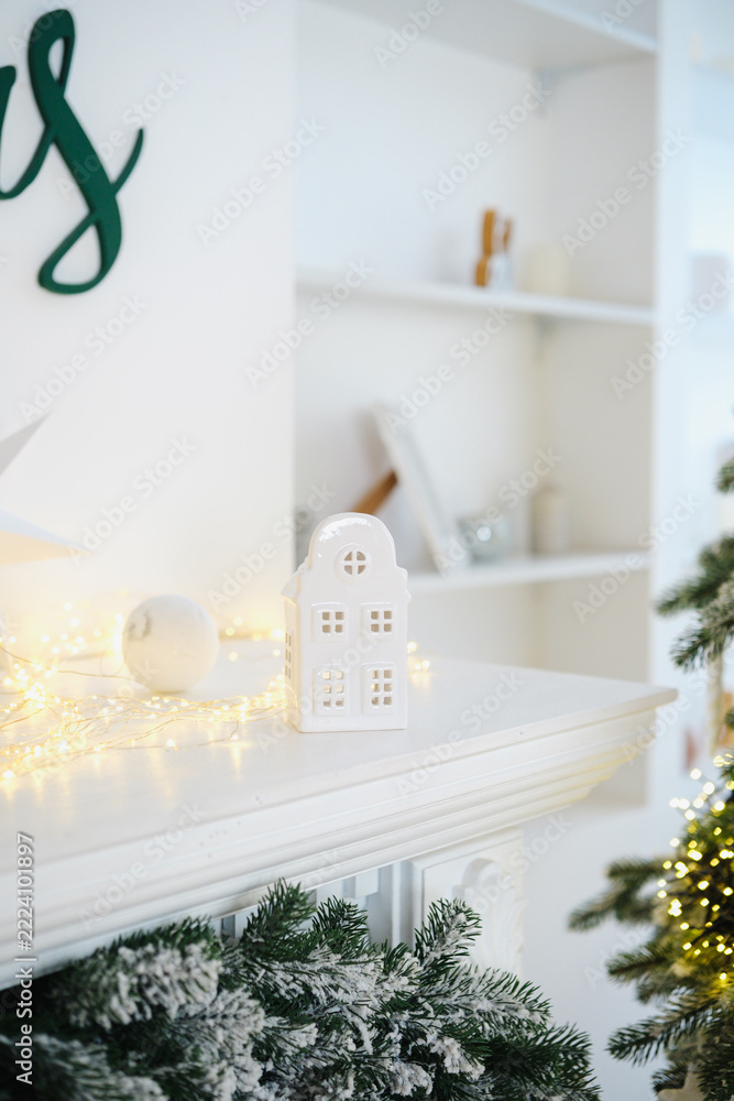 Naklejka premium White festive fireplace mantel with miniature house, glowing lights, green garland, and snowy tree.