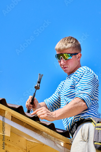 Bitumen roofing sheets being secured to wooden roof battens by worker using hammer to drive nail through corrugated roofing material.