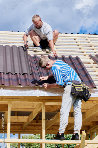 Asphalt roofing sheets are aligned by two construction workers fitting corrugated panels onto timber roof battens during house construction.