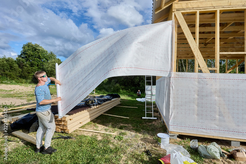 House wrap being unrolled by builder beside timber frame wall during installation of protective membrane over wooden studs and braces.