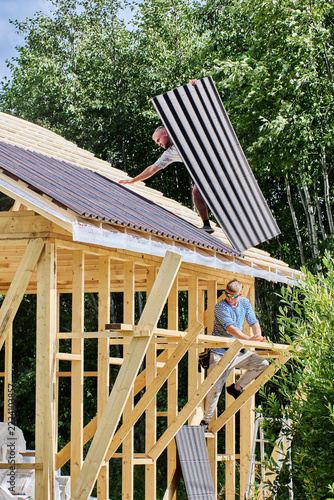 Asphalt roofing sheets are passed to worker on sloped timber roof while another builder handles panels during installation of roofing.