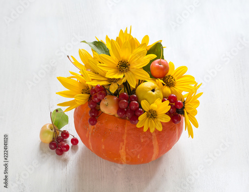 A beautiful autumn arrangement of bright yellow Topinambur flowers,  small Apples, and red Viburnum berries, placed inside an orange pumpkin against a white wooden background.