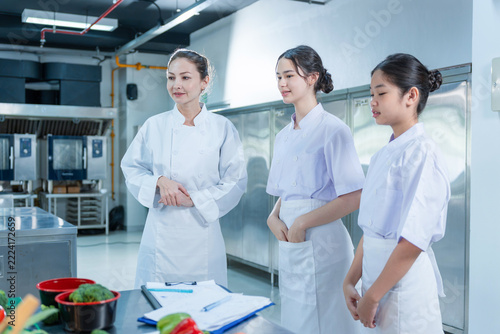 Professional chef giving a lecture to young female students in culinary school training class, Head chef mentor standing with student chefs during training in modern restaurant kitchen