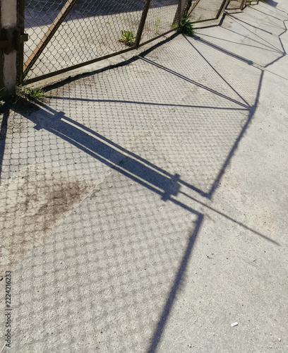 Sunlight casts the silhouette of a metal security fence onto an asphalt path.