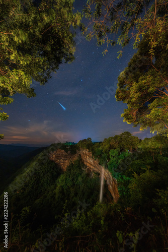 Tad Mok Waterfall and cliff under a starry night sky. A bright meteor streaking above.