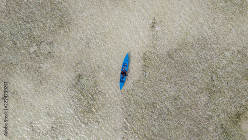 Aerial view of a person paddling a blue kayak through the shallow, crystal-clear turquoise waters of a coral reef in Wakatobi, Southeast Sulawesi, Indonesia.