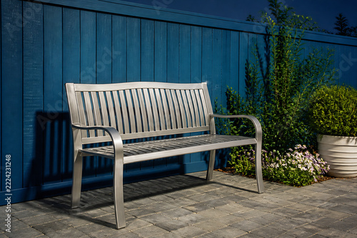 A modern metal park bench is situated on a paved patio against a dark blue wooden fence with greenery in the background.