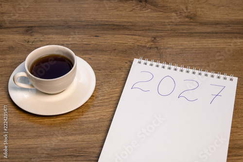 A cup of black coffee beside a notepad marked 2027 on a wooden table. New Year preparation and planning routine concept.