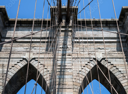 2019 New York City, USA; Architectural detail of the stone tower of the Brooklyn Bridge with gothic arches, steel suspension cables, year 1875 plaque