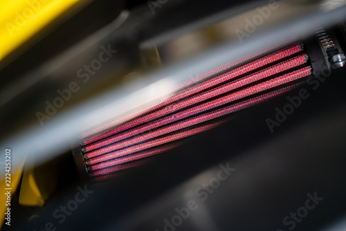 Close-up of a red performance engine air filter, photographed through the cooling vent grille on the hood of a sports car.