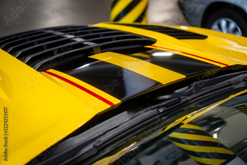 Close-up of glossy black racing stripes with red accents on the yellow hood of a sports car, featuring a custom vinyl wrap and air vents.