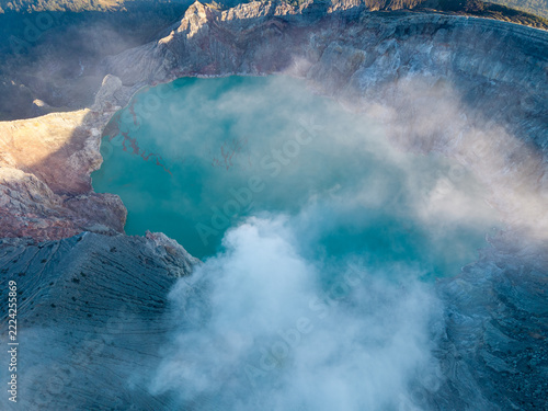 Clouds drift over a turquoise volcanic crater lake. Rugged, gray slopes encircle the glowing water at Mount Ijen