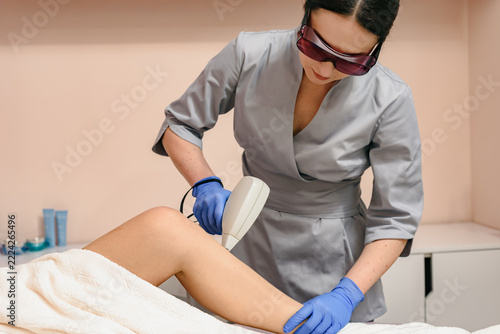 Female clinician wearing protective goggles and blue gloves uses handheld laser device for hair removal on client's exposed lower leg while client rests on treatment bed inside clean beauty room