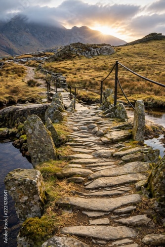 A rustic stone pathway crosses a small river, surrounded by golden grass and hills, with misty mountains under a glowing sunset in the distance.