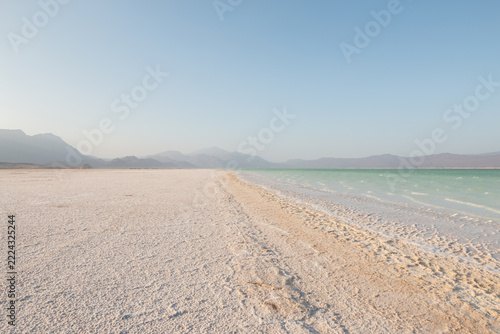 Salt flat meeting turquoise lake with hazy mountains under clear blue sky, Lake Assal, Djibouti