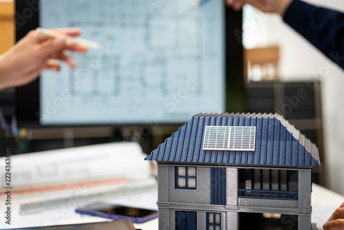 Obraz na plátně Miniature house with rooftop solar panels on architectural plans, with architects reviewing a home design in the background
