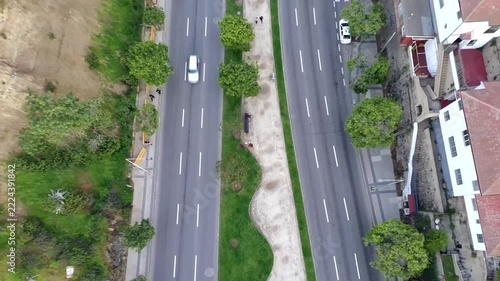 Top down drone footage of a wide urban avenue with moving cars, multiple traffic lanes, trees and a landscaped central median. Aerial perspective of city roads and transportation infrastructure.