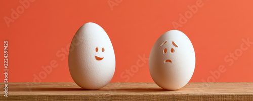 Two white eggs with happy and sad faces drawn on them stand on wooden surface against orange background, expressing contrasting emotions