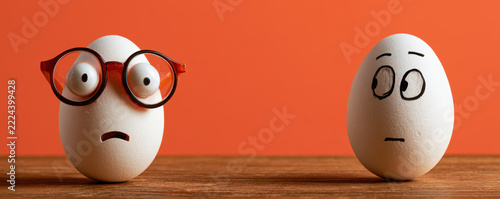 Two eggs with drawn faces showing surprised and worried emotions, one wearing glasses, on wooden surface with orange background