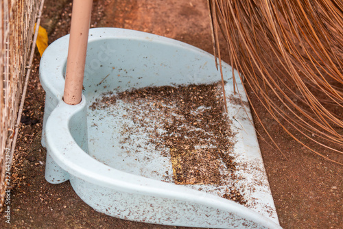 Light blue plastic dustpan filled with dirt, seed husks and dust beside a natural palm fiber broom and a pink handle on a reddish dirt ground outdoors