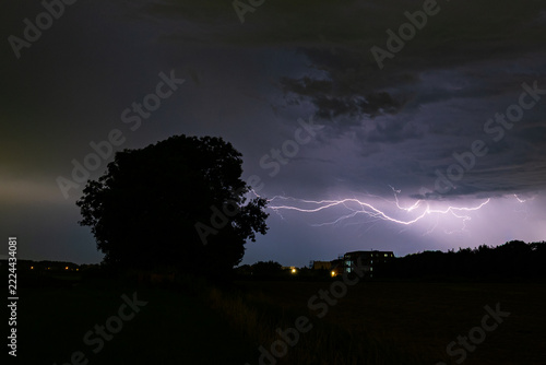 Dramatic lightning storm over rural nightscape with tree silhouette and distant buildings at night.
