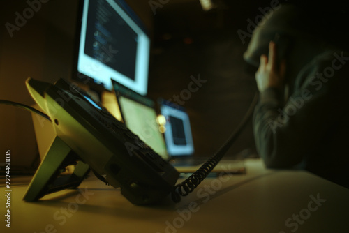 Defocused Hacker in Dark Hoodie Making Telephone Call Among Computer Screens. Telephone Fraud Concept.