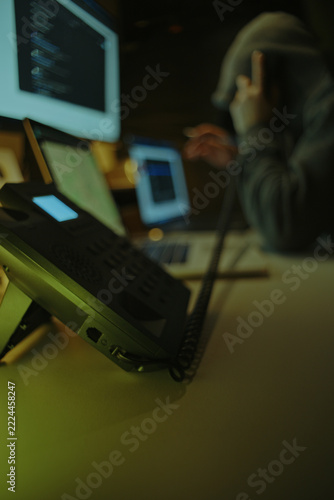 Defocused Hacker in Dark Hoodie Making Telephone Call Among Computer Screens. Telephone Fraud Concept.