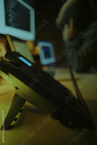 Defocused Hacker in Dark Hoodie Making Telephone Call Among Computer Screens. Telephone Fraud Concept.