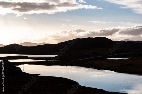 Silhouetted hills meet calm waters under a cloudy sky. Sunlight glows behind distant peaks, casting soft tones. Reflections ripple gently, mirroring the serene landscape