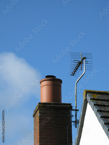 Clear sky with soft cloud above chimney and antenna. Red brick chimney stands beside metal antenna. Antenna leans slightly, mounted on metal bracket. Image conveys calm. Use for home, weather