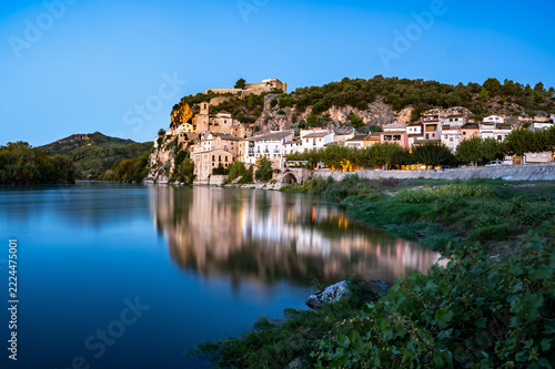   A picturesque historic village with an ancient fortress nestled atop a steep rocky cliff, perfectly reflected in the calm waters of a wide river under a bright blue sky  