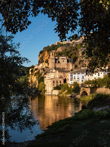   A picturesque historic village with an ancient fortress nestled atop a steep rocky cliff, perfectly reflected in the calm waters of a wide river under a bright blue sky  